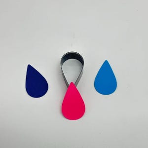 Könnte beinhalten: Ein tropfenförmiger Ohrringausstecher mit drei tropfenförmigen Teilen in Blau und Pink. Der Ausstecher ist schwarz und die Tropfen sind blau und pink.