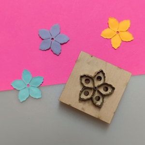 Può includere: Un timbro in legno a forma di fiore con un contorno metallico, accanto a tre fiori di carta colorati in blu, viola e giallo. Lo sfondo è rosa e grigio.