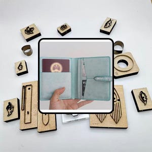 May include: A light blue leather wallet with a snap closure, open to reveal a passport and cards. Surrounding the wallet are various wooden stamps and metal rings, likely for crafting or leatherwork. The stamps feature leaf and geometric designs.