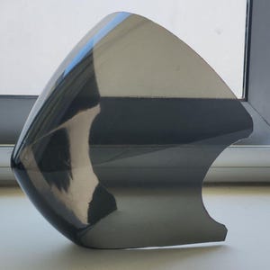 May include: A smooth, curved, semi-transparent dark gray plastic motorcycle windshield or fairing component. The surface is highly reflective, showing light reflections and a glossy finish with a distinct pointed top and a contoured cutout edge.