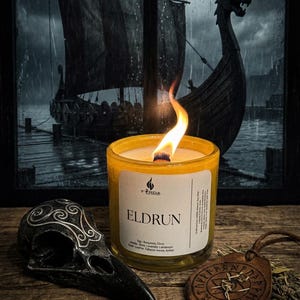 May include: A lit yellow candle in a glass jar with the word "ELDRUN" on the label. A black raven skull and a leather pendant with runes are in the foreground. A Viking ship is visible in the background.