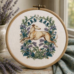 May include: A circular wooden embroidery hoop containing a cross-stitch design of a tan greyhound in a running pose, surrounded by a detailed wreath of blue and purple flowers, green leaves, and small orange berries on a white fabric background.