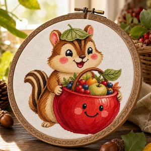 May include: A circular wooden embroidery hoop containing a cross-stitch design of a brown chipmunk with a green leaf on its head, holding a large red basket with a smiling face that is filled with various colourful fruits and berries.