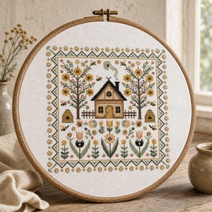 May include: A circular wooden embroidery hoop containing a cross-stitch pattern on white fabric. The design features a central house with a chimney, flanked by two trees, beehives, and various flowers in shades of yellow, green, and brown.