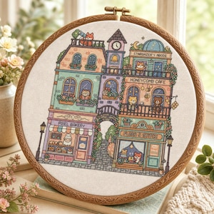 Może przedstawiać: Okrągły drewniany tamborek z haftem krzyżykowym przedstawiającym kompleks budynków. Scena zawiera Pixel Bakery, Honeycomb Cafe, sklep Flora & Stitch oraz Stargazer's Abode, zamieszkane przez różne małe postacie zwierząt widoczne w oknach.