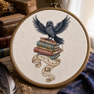 May include: A circular embroidery hoop containing a cross-stitch design of a black crow perched atop a stack of four colorful books. Below the books is a beige scroll featuring celestial symbols, stars, and a small open book illustration.