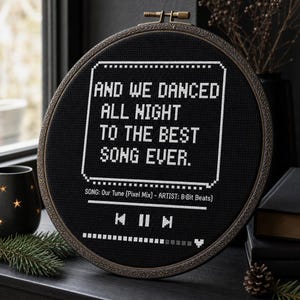 May include: A circular embroidery hoop containing black fabric with white cross-stitched text that reads: AND WE DANCED ALL NIGHT TO THE BEST SONG EVER. Below this, it displays SONG: Our Tune (Pixel Mix) - ARTIST: 8-Bit Beats and media controls.