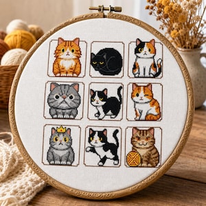 May include: A circular wooden embroidery hoop containing a white fabric panel with a cross-stitch pattern of nine distinct cats. The cats are arranged in a grid, featuring various colours and poses, including a cat wearing a small yellow crown.