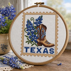 May include: A circular wooden embroidery hoop containing a cross-stitch design of a brown cowboy boot and blue lupine flowers, framed by a tan postage stamp border. The word TEXAS is stitched in blue block letters at the bottom of the design.
