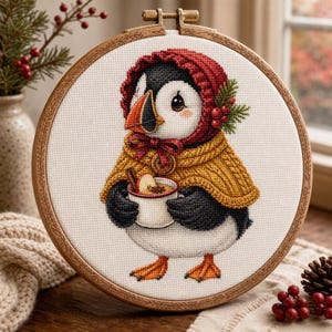 Puede incluir: Un bastidor de bordado circular de madera que contiene un diseño de punto de cruz de un frailecillo con un gorro rojo y una capa amarilla. El frailecillo sostiene una taza blanca llena de una bebida con una rama de canela y anís estrellado.