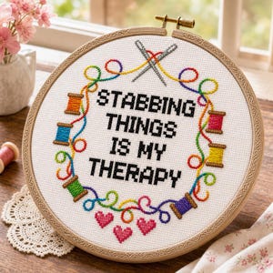 May include: A circular wooden embroidery hoop containing white fabric with the black stitched text STABBING THINGS IS MY THERAPY. The design features a colorful rainbow thread border, small spools of thread, two crossed needles, and three pink hearts.