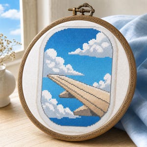 May include: A cross-stitch embroidery piece framed in a round, ornate bronze-coloured hoop. The design depicts an aeroplane window view showing a tan aeroplane wing against a bright blue sky filled with white, pixelated-style clouds.