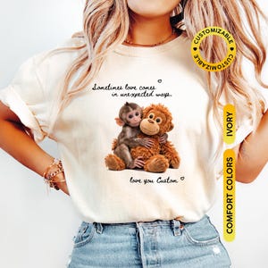 May include: Ivory-colored t-shirt featuring a graphic of two hugging monkeys, with the text "Sometimes love comes in unexpected ways." and "love you Custom." The shirt also has a "Customizable" logo and "Comfort Colors" label.