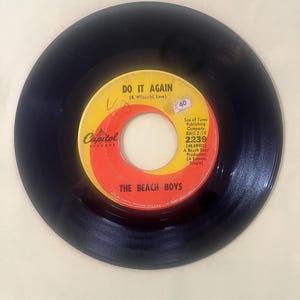 Könnte beinhalten: Eine schwarze Schallplatte mit einem gelb-roten Label. Das Label zeigt den Text "DO IT AGAIN" und "THE BEACH BOYS". Das Capitol Records-Logo ist sichtbar. Die Schallplatte ist eine 45-RPM-Single.