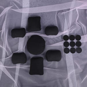 May include: A collection of black foam pads and circular stickers arranged on a white, sheer fabric. The pads vary in shape, including rectangular, square, and rounded forms. The stickers are in a grid of three rows and three columns.
