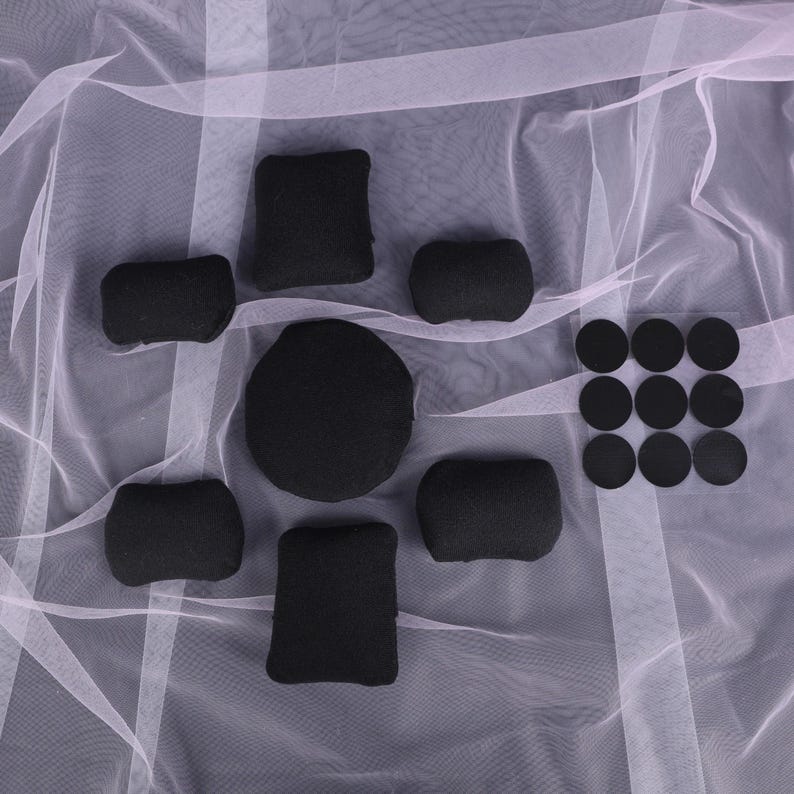 May include: A collection of black foam pads and circular stickers arranged on a white, sheer fabric. The pads vary in shape, including rectangular, square, and rounded forms. The stickers are in a grid of three rows and three columns.