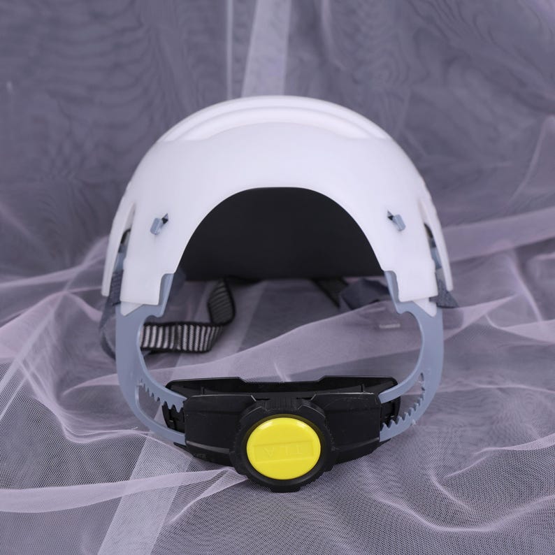 May include: White safety helmet with a black and yellow adjustment dial. The helmet has a gray suspension system and a black and white adjustable strap. The helmet is designed to protect the head from impact.
