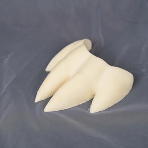 May include: A three-pronged, off-white, claw-like object with a smooth, matte texture. The piece features three distinct, pointed extensions that taper to rounded tips, connected at a central base, resembling a stylized animal or creature claw.