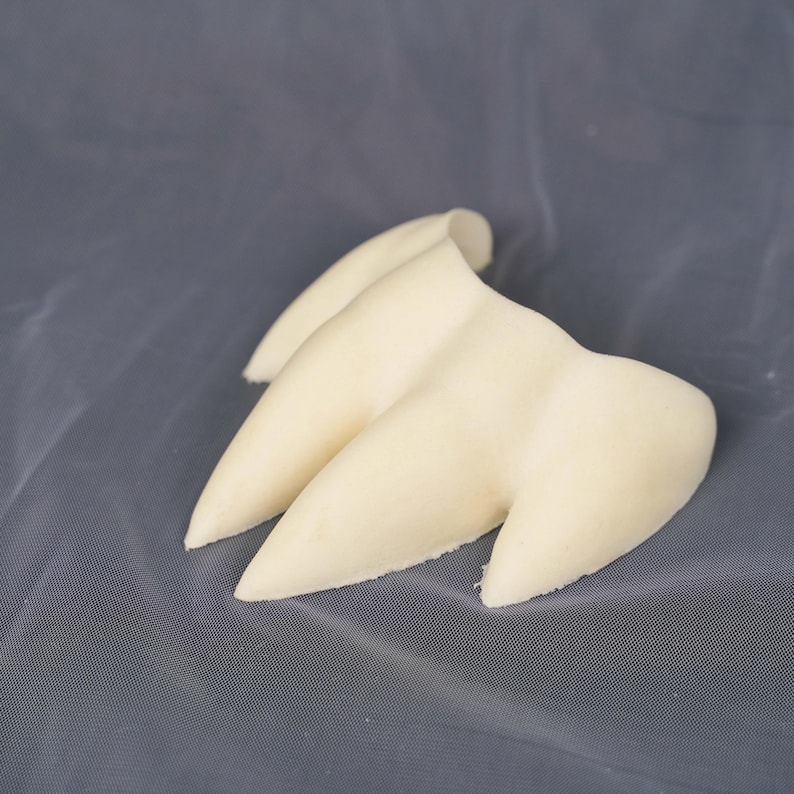 May include: A three-pronged, off-white, claw-like object with a smooth, matte texture. The piece features three distinct, pointed extensions that taper to rounded tips, connected at a central base, resembling a stylized animal or creature claw.