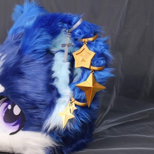 Peut inclure: Une tête en fourrure bleue et blanche avec de grands yeux violets et blancs. Des breloques dorées en forme d'étoile et une croix argentée pendent de la tête. La tête fait probablement partie d'un costume ou d'un cosplay.