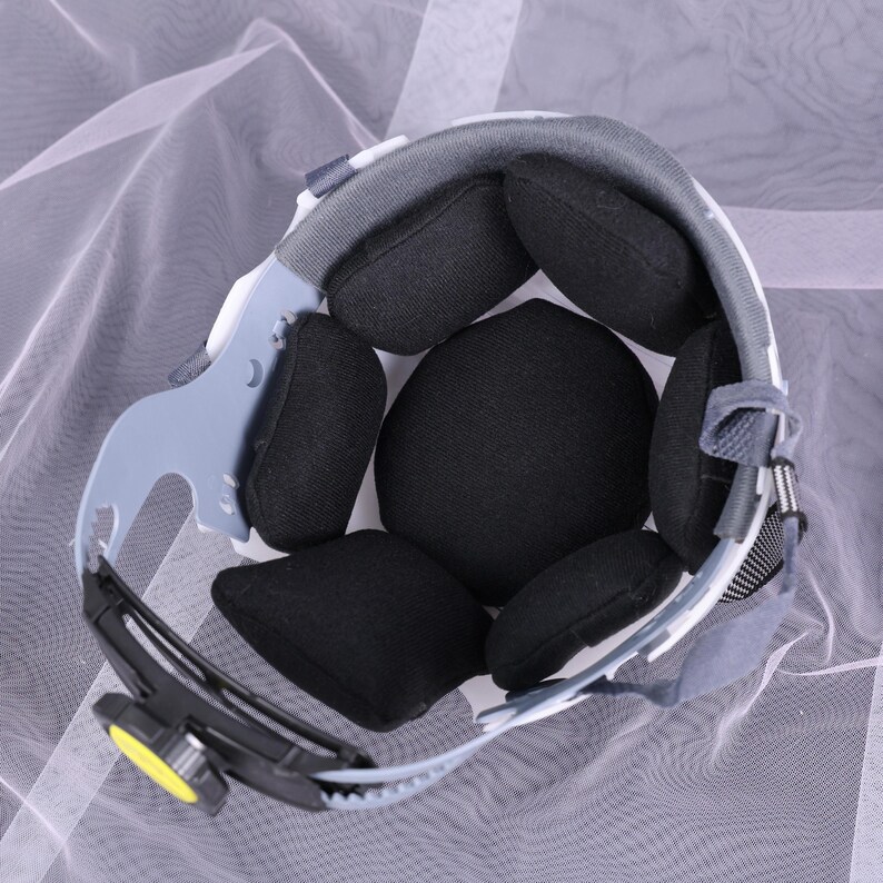 May include: Interior view of a safety helmet, showcasing its internal components. The helmet features a white and gray outer shell, with black padded cushions for comfort and impact absorption. A gray suspension system and a yellow adjustment knob are visible.