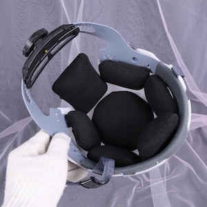 May include: An inside view of a white safety helmet with black padding. The helmet has a gray adjustable head support and a black adjustment knob. The interior features several black, cushioned pads for comfort and protection.