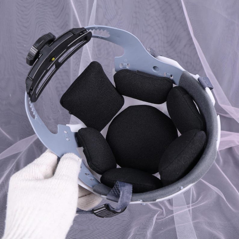 May include: An inside view of a white safety helmet with black padding. The helmet has a gray adjustable head support and a black adjustment knob. The interior features several black, cushioned pads for comfort and protection.