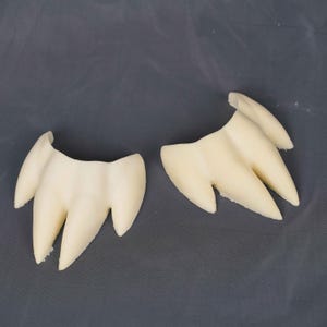 May include: Two off-white, three-pronged claw-like objects with smooth, curved surfaces. Each piece features a central point flanked by two smaller, tapered points, resembling a stylized animal paw or tooth structure with a matte finish.