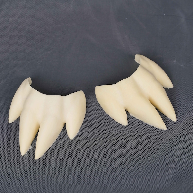 May include: Two off-white, three-pronged claw-like objects with smooth, curved surfaces. Each piece features a central point flanked by two smaller, tapered points, resembling a stylized animal paw or tooth structure with a matte finish.