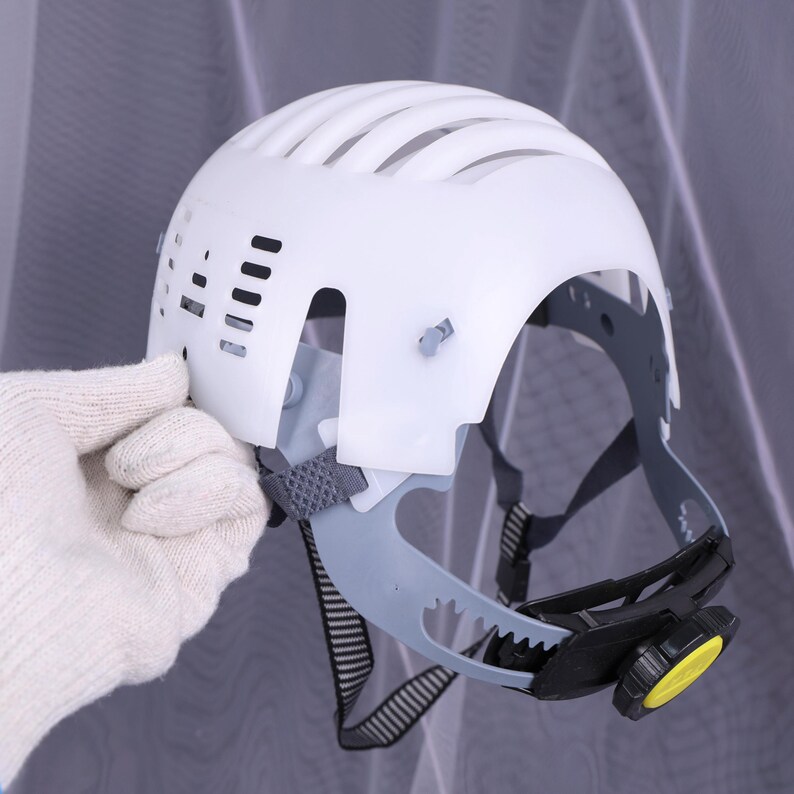 May include: White safety helmet with a ribbed top and ventilation slots. The helmet has a gray interior harness and a black and yellow adjustment knob. A gloved hand is holding the helmet.