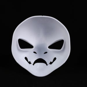 May include: A plain white mask with a simplified face design. It has angular eye cutouts, small holes for nostrils, and a curved mouth with jagged edges. The mask is made of a smooth, solid material.