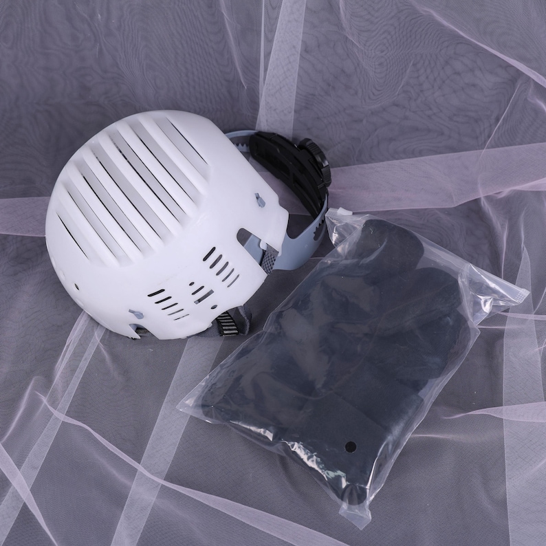 May include: A white helmet with black accents and a clear plastic bag containing black items. The helmet has a series of vertical vents and a strap. The items in the bag are black and appear to be protective gear.