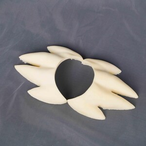 May include: A cream-colored, rigid, molded plastic hair accessory featuring a stylized, spiky, multi-pointed design that radiates outward from a central circular opening. The piece has a smooth, matte finish and a symmetrical, fan-like structure.