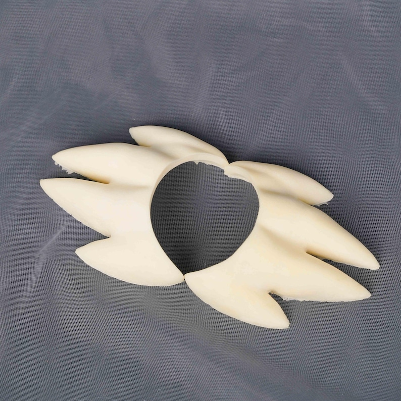May include: A cream-colored, rigid, molded plastic hair accessory featuring a stylized, spiky, multi-pointed design that radiates outward from a central circular opening. The piece has a smooth, matte finish and a symmetrical, fan-like structure.