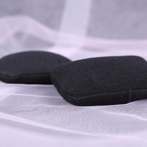 May include: Two small, black, fabric-covered objects, one oval and one square, rest on a white, sheer, textured surface. The objects appear to be small pillows or pads, possibly for jewelry or display.