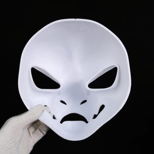 May include: A white plastic mask with angular eye openings, small nose holes, and a curved mouth. The mask is held by a gloved hand against a black background. The mask's design suggests a theatrical or costume purpose.