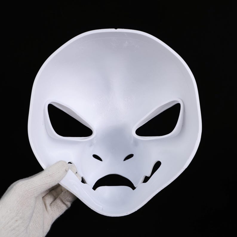 May include: A white plastic mask with angular eye openings, small nose holes, and a curved mouth. The mask is held by a gloved hand against a black background. The mask's design suggests a theatrical or costume purpose.