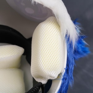 May include: A helmet interior featuring cream-colored mesh padding cushions attached to the inner shell. The exterior of the helmet is covered in long, vibrant blue faux fur, with a section of white faux fur visible along the top edge.