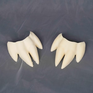 May include: Two off-white, molded plastic dental prosthetics, each featuring three pointed, fang-like protrusions connected by a curved base. The items have a smooth, matte finish and are positioned side-by-side against a neutral gray background.