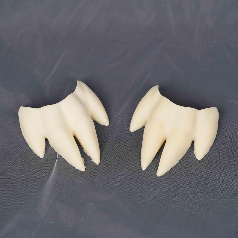 May include: Two off-white, molded plastic dental prosthetics, each featuring three pointed, fang-like protrusions connected by a curved base. The items have a smooth, matte finish and are positioned side-by-side against a neutral gray background.