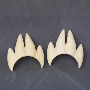 May include: Two off-white, three-pronged claw-shaped foam pieces with a curved base. Each piece features a central split between the two longest points and two shorter side points, creating a symmetrical, pointed design with a smooth texture.
