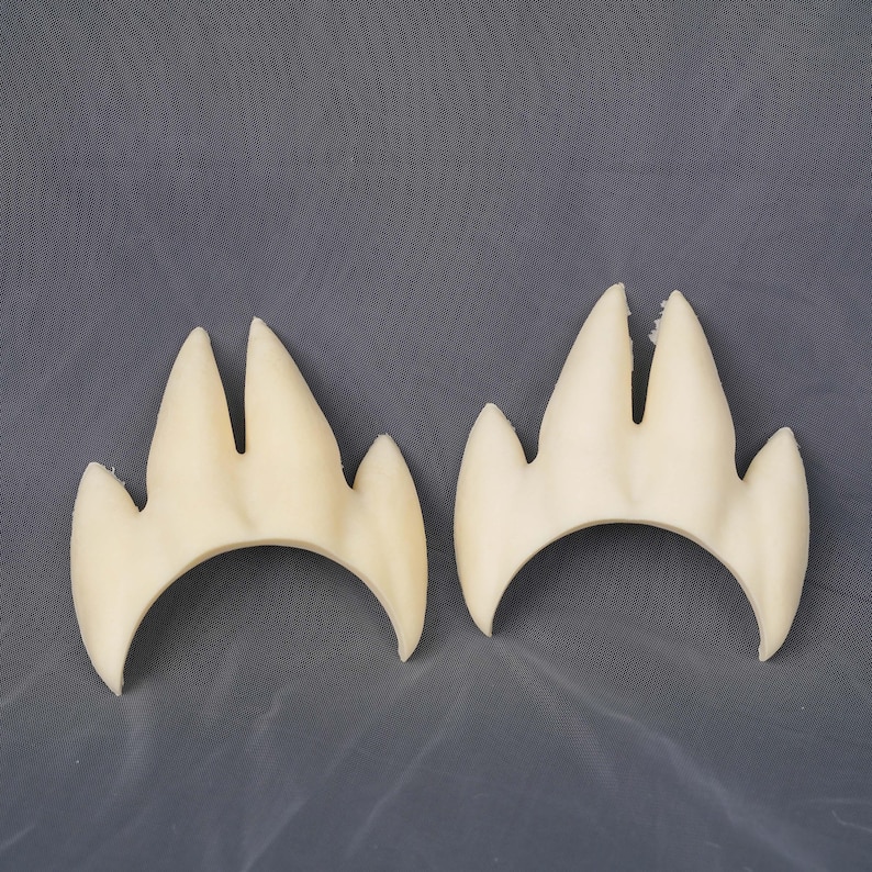 May include: Two off-white, three-pronged claw-shaped foam pieces with a curved base. Each piece features a central split between the two longest points and two shorter side points, creating a symmetrical, pointed design with a smooth texture.