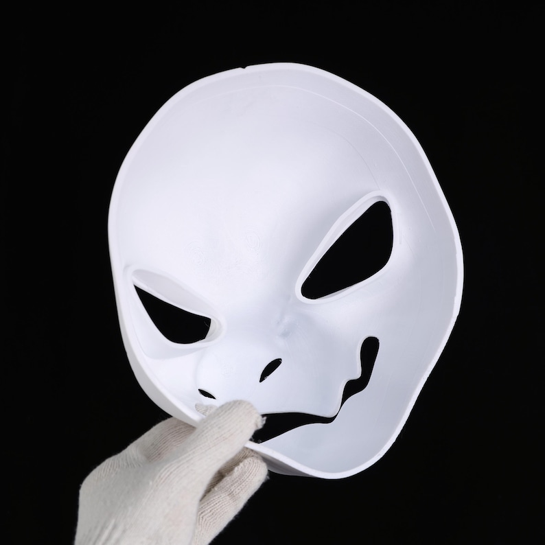 May include: A white plastic mask with large eye openings and a stylized mouth. The mask is held by a gloved hand against a black background. The mask's design suggests a character or costume piece.