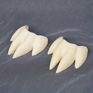 May include: Two off-white, three-pronged claw-shaped objects with a smooth, matte finish. Each piece features three distinct, pointed extensions connected at a curved base, resembling a stylized animal paw or talon design in a neutral color.