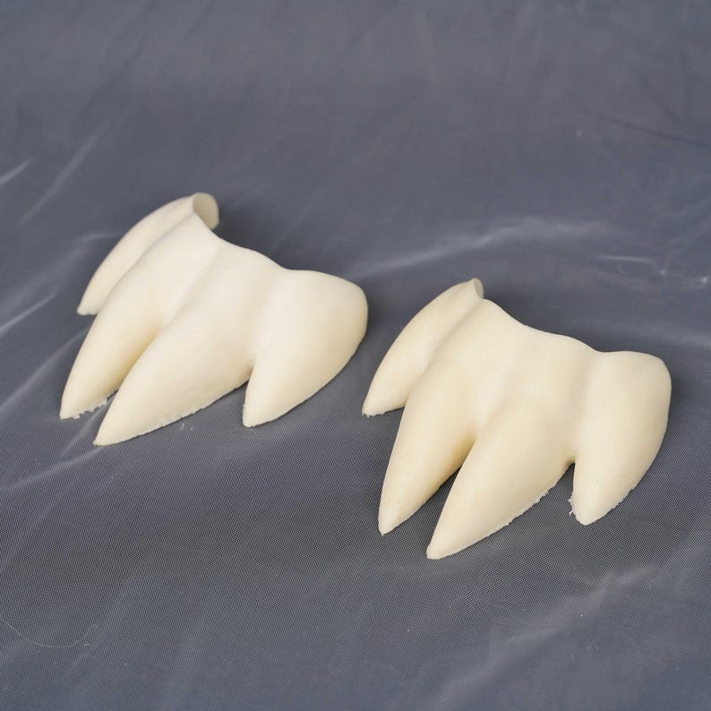 May include: Two off-white, three-pronged claw-shaped objects with a smooth, matte finish. Each piece features three distinct, pointed extensions connected at a curved base, resembling a stylized animal paw or talon design in a neutral color.