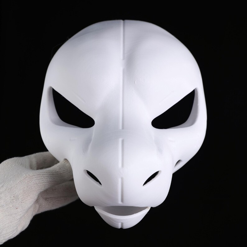 May include: A white mask with a dragon-like design. The mask has large, angled eye openings, small nostril holes, and a slightly open mouth. The mask is held by a gloved hand against a black background.
