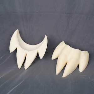 May include: Two off-white, foam-like prosthetic pieces shaped like sets of pointed fangs or teeth. One piece is curved and hollowed out on the interior, while the other is a solid, three-pronged structure with a smooth, matte surface texture.