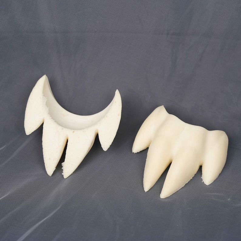 May include: Two off-white, foam-like prosthetic pieces shaped like sets of pointed fangs or teeth. One piece is curved and hollowed out on the interior, while the other is a solid, three-pronged structure with a smooth, matte surface texture.