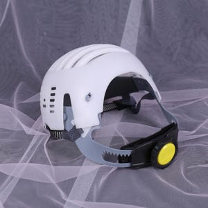 May include: A white safety helmet with a curved design and ventilation slots. The helmet has a gray and black adjustable chin strap with a yellow dial. The helmet is designed for head protection.