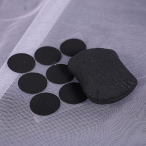 May include: A collection of black circular fabric patches and a small, black rectangular fabric object. The items are arranged on a sheer, white fabric background. The patches are evenly spaced, and the rectangular object is slightly rounded.